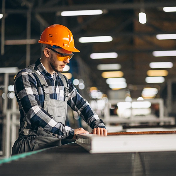 A person in a plaid shirt and orange hard hat is leaning on a workbench in an industrial setting, with their face obscured for privacy. The environment suggests manufacturing or assembly work.